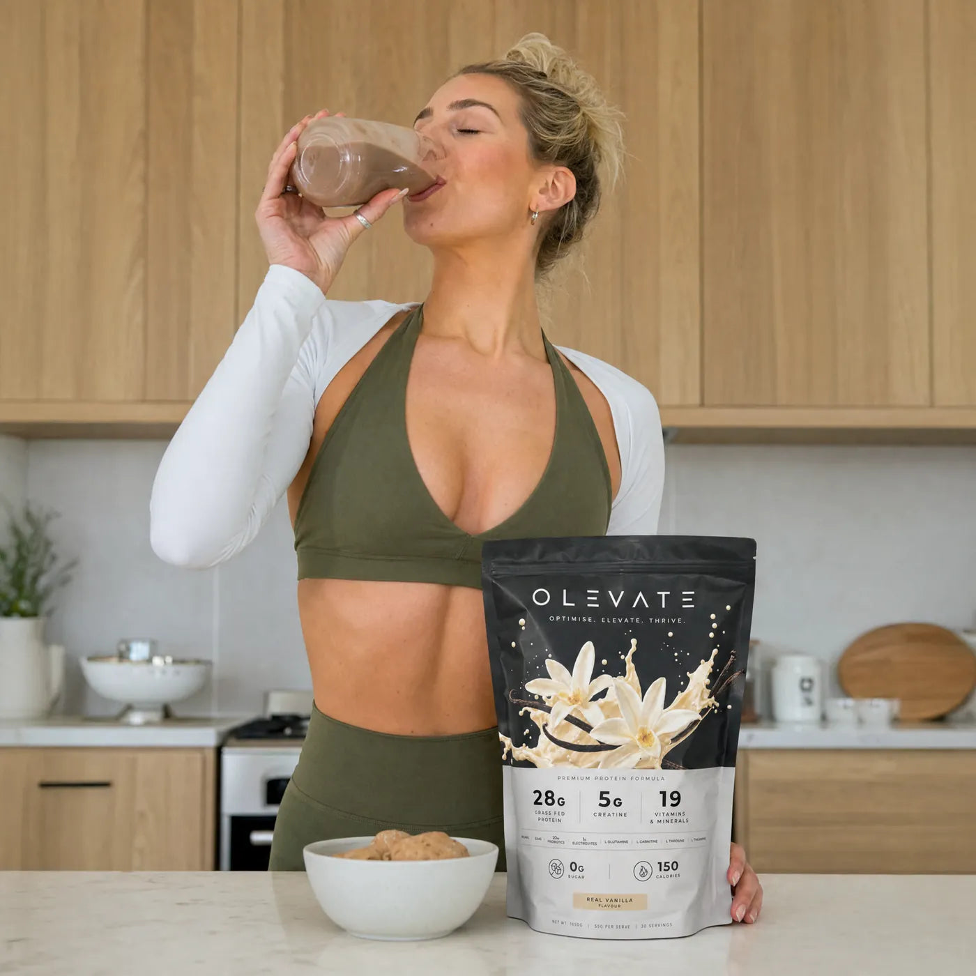 A person in a kitchen drinking an OLEVATE shake beside the Real Vanilla pouch